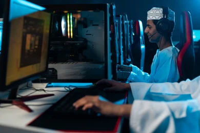 شاب عربي خليجي عماني يرتدي دشداشة بيضاء و كمة مطرزة يجلس بجانب صديقه داخل مركز الالعاب الالكترونية , انشطة شبابية ممتعة , الجلوس امام الحاسب المكتبي , النقر على لوحة المفاتيح