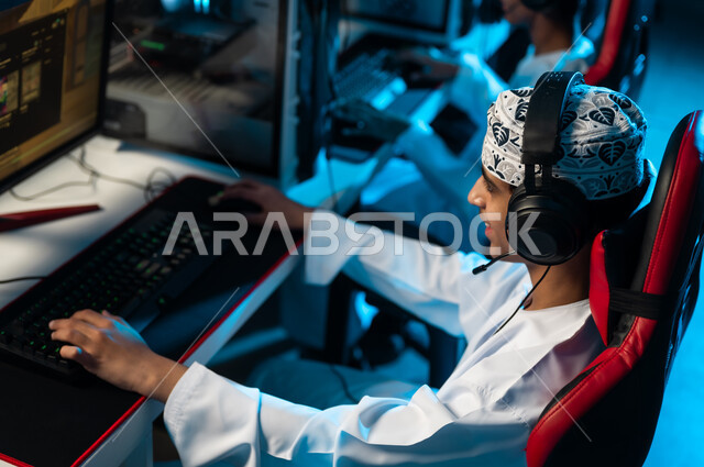 اللعب بحماس و تركيز داخل المركز الالكتروني للالعاب , شاب عربي خليجي عماني يرتدي دشداشة بيضاء و كمة مطرزة يضع سماعة رأس اثناء اللعب للتواصل مع الاصدقاء , الجلوس امام الجهاز التقني الحديث