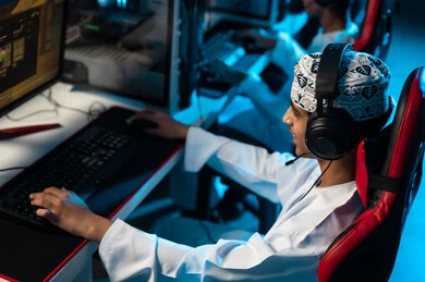 اللعب بحماس و تركيز داخل المركز الالكتروني للالعاب , شاب عربي خليجي عماني يرتدي دشداشة بيضاء و كمة مطرزة يضع سماعة رأس اثناء اللعب للتواصل مع الاصدقاء , الجلوس امام الجهاز التقني الحديث