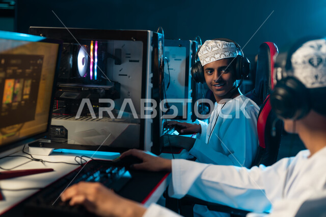 شاب عربي خليجي عماني يرتدي دشداشة بيضاء و كمة مطرزة يشارك صديقه اللعب الالكتروني , الجلوس على مقاعد جلدية حمراء , اوقات ممتعة بين الاصدقاء , وضع سماعة الرأس لتسهيل التواصل مع اللاعبين