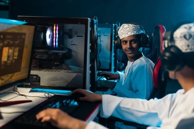 A young Arab Gulf Omani man wearing a white dishdasha and an embroidered kumma is playing video games with his friend, sitting on red leather chairs, enjoying fun times with friends, wearing headphones to facilitate communication with the players.