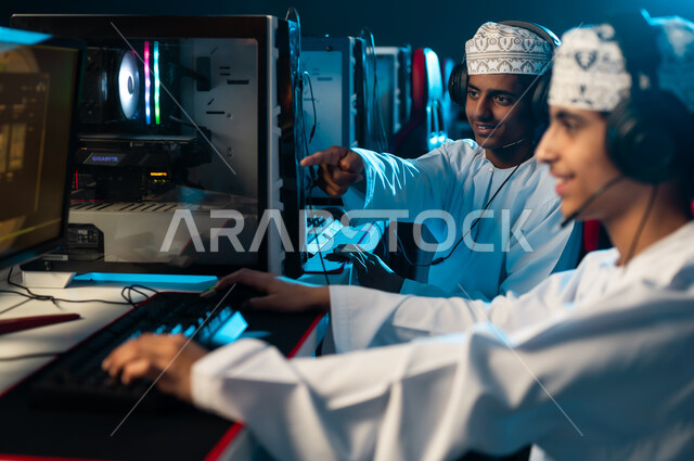 مشاركة الالعاب الحماسية المثيرة مع الاصدقاء , شابان عربيان خليجيان عمانيان يرتديان الدشداشة الييضاء و الكمة المطرزة يجلسان امام الحاسب المكتبي , الاشارة باصبع السبابة الي الشاشة, ايماءات الفرحة و السعادة