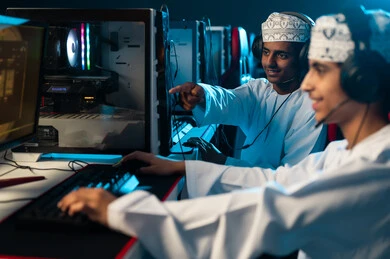 مشاركة الالعاب الحماسية المثيرة مع الاصدقاء , شابان عربيان خليجيان عمانيان يرتديان الدشداشة الييضاء و الكمة المطرزة يجلسان امام الحاسب المكتبي , الاشارة باصبع السبابة الي الشاشة, ايماءات الفرحة و السعادة