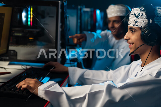النظر الي شاشة الحاسب المكتبي بسعادة , شابان عربيان خليجيان عمانيان يرتديان الدشداشة البيضاء و الكمة المطرزة يجلسان على المقاعد الجلدية الحمراء , الاشارة باصبع السبابة الى الشاشة
