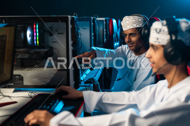 الاشارة باصبع السبابة الى شاشة الحاسب المكتبي , شابان عربيان خليجيان عمانيان يرتديان دشداشة بيضاء و كمة مطرزة يجلسان امام الجهاز التقني , العاب الكترونية حماسية بين الاصدقاء