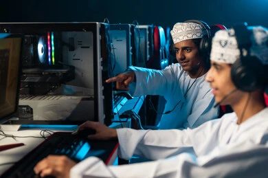 الاشارة باصبع السبابة الى شاشة الحاسب المكتبي , شابان عربيان خليجيان عمانيان يرتديان دشداشة بيضاء و كمة مطرزة يجلسان امام الجهاز التقني , العاب الكترونية حماسية بين الاصدقاء
