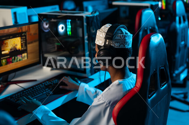 A young Arab Gulf Omani man wearing a white dishdasha and an embroidered kumma sits in front of a desktop computer, enjoying electronic games with friends, with a background image, sitting on a red leather chair.