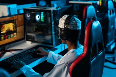 شاب عربي خليجي عماني يرتدي دشداشة بيضاءو كمة مطرزة يجلس امام الحاسب المكتبي ,العاب الكترونية ممتعة مع الاصدقاء , صورة خلفية , الجلوس على مقعد جلدي احمر