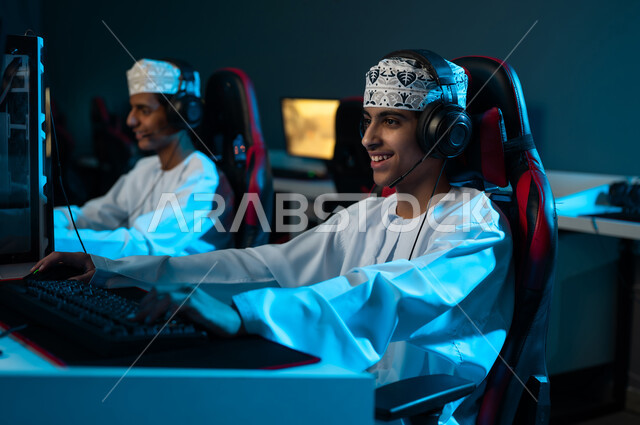 النظر الي الشاشة بابتسامةو فرحة , شابان عربيان خليجيان عمانيان يرتديان الدشداشة البيضاء و الكمة المطرزة يضعان سماعة رأس لسهولة التواصل اثناء اللعب , العاب الكترونية جماعية بين الاصدقاء