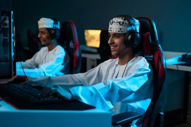 النظر الي الشاشة بابتسامةو فرحة , شابان عربيان خليجيان عمانيان يرتديان الدشداشة البيضاء و الكمة المطرزة يضعان سماعة رأس لسهولة التواصل اثناء اللعب , العاب الكترونية جماعية بين الاصدقاء