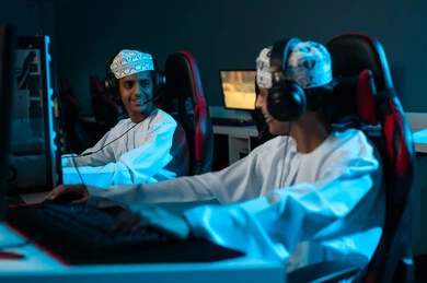 اللعب الالكتروني بين الاصدقاء , شابان عربيان خليجيان عمانيان يرتديان الكمة المطرزة و الدشداشة البيضاء يجلسان على المقاعد الجلدية الحمراء , وضع سماعات الرأس لسهولة التواصل اثناء اللعب , ايماءات السعادة و الابتسامة