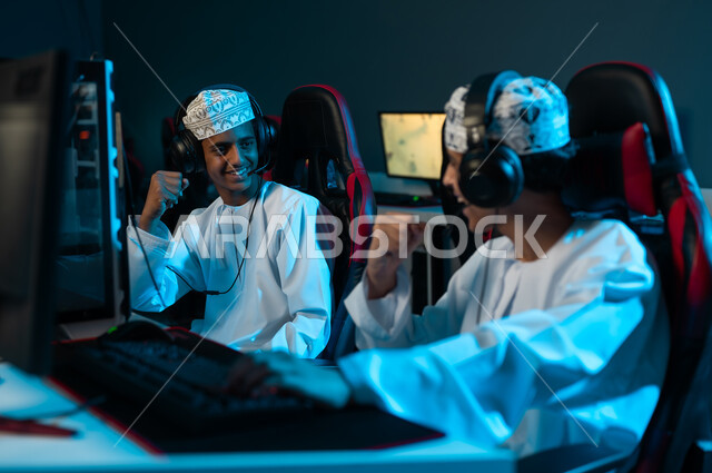 Two young Omani Arab Gulf men wearing white dishdasha and embroidered kumma are looking at each other happily, raising their fists in the air as a sign of victory, gestures of joy and happiness.