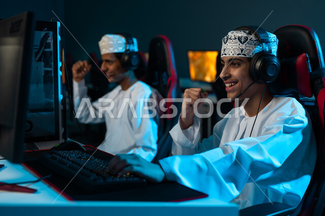 رفع قبضة اليد لاعلى علامة على تحقيق الفوز , شابان عربيان خليجيان عمانيان يرتديان الدشداشة البيضاء و الكمة المطرزة يلعبان الالعاب الالكترونية الجماعية , ايماءات السعادة و الفرحة