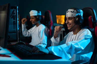 رفع قبضة اليد لاعلى علامة على تحقيق الفوز , شابان عربيان خليجيان عمانيان يرتديان الدشداشة البيضاء و الكمة المطرزة يلعبان الالعاب الالكترونية الجماعية , ايماءات السعادة و الفرحة