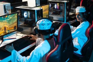Two young Arab Gulf Omanis wearing white dishdasha and embroidered kumma are looking at each other while playing video games on a desktop computer, with gestures of happiness and smiles.
