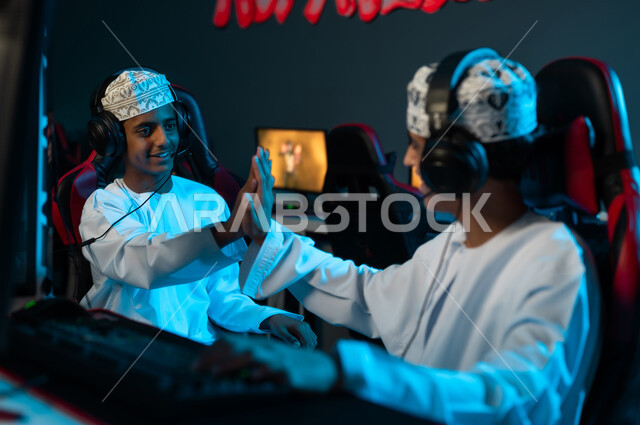 ضرب الكفين في الاعلى , شابان عربيان خليجيان عمانيان يجلسان امام جهاز الحاسب المكتبي , ايماءات السعادة و الابتسامة علامة على تحقيق الفوز , اوقات مميزة بين الاصدقاء
