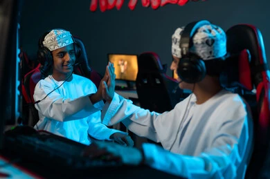 ضرب الكفين في الاعلى , شابان عربيان خليجيان عمانيان يجلسان امام جهاز الحاسب المكتبي , ايماءات السعادة و الابتسامة علامة على تحقيق الفوز , اوقات مميزة بين الاصدقاء