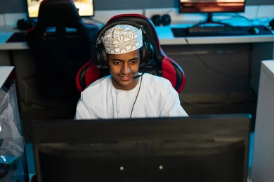 Exciting various activities at the electronic games center, a young Arab Gulf Omani man wearing a white dishdasha and an embroidered kumma sits on a red leather chair, spending enjoyable times with friends.