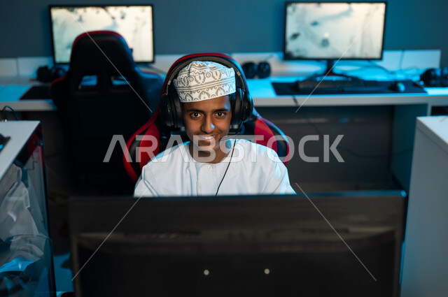 شاب عربي خليجي عماني يرتدي الكمة المطرزة و الدشداشة البيضاء يجلس امام شاشة الحاسب المكتبى , النظر الي الكاميرا بابتسامة , صورة مقربة , وضع سماعة الرأس لسماع الاصدقاء اثناء اللعب