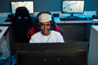 A young Omani Gulf Arab man wearing an embroidered kumma and a white dishdasha sits in front of a desktop computer screen, looking at the camera with a smile, in a close-up shot, wearing headphones to listen to friends while playing.