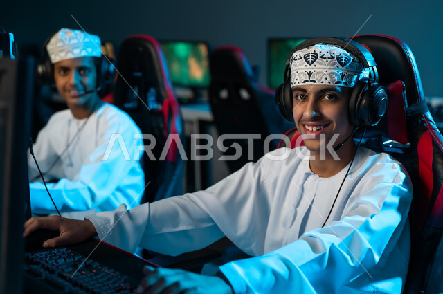 Smiling at the camera, two young Arab Gulf Omanis wearing white dishdasha and embroidered kumma are sitting in front of a desktop computer, playing multiplayer electronic games among friends.