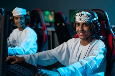 نظرات الى الكاميرا بابتسامة , شابان عربيان خليجيان عمانيان يرتديان الدشداشة البيضاء و الكمة المطرزة يجلسان امام الحاسب المكتبي , العاب الكترونية جماعية بين الاصدقاء