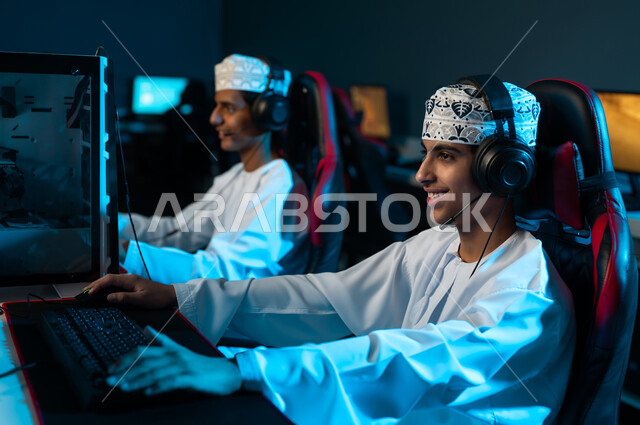 Focusing on the computer screen, two young Arab Gulf Omanis wearing white dishdasha and embroidered kumma are sitting on leather seats, the ease of communication and playing with friends remotely.