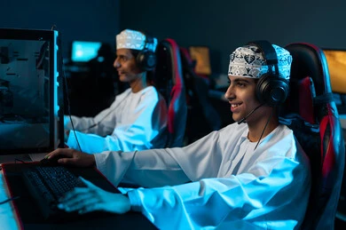 النظر الى شاشة الحاسب بتركيز , شابان عربيان خليجيان عمانيان يرتديان دشداشة بيضاء و كمة مطرزة يجلسان على المقاعد الجلدية , سهولة التواصل و اللعب مع الاصدقاء عن بعد