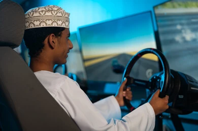 شاب عربي خليجي عماني صغير ينظر الي شاشة السباق بتركيز و حماس , صورة خلفية الامساك بعجلة القيادة بثبات , انشطة شبابية ممتعة , مركز الالعاب الالكترونية