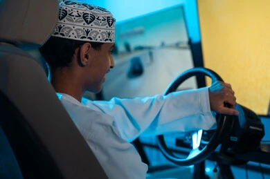 النظر الي الشاشة المعلقة بفرحة و ابتسامة , شاب عربي خليجي عماني يرتدي الدشداشة البيضاء و الكمة المطرزة يمسك مقود السيارة , التنافس و الحماس بين الاصدقاء , صورة خلفية