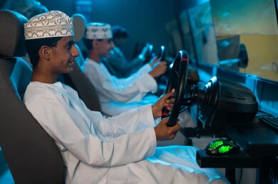 A group of young Arab Gulf Omanis wearing white dishdashas and embroidered kumas, sitting in front of screens playing car racing games, engaging in collective youth activities, looking with focus and professionalism to achieve victory.
