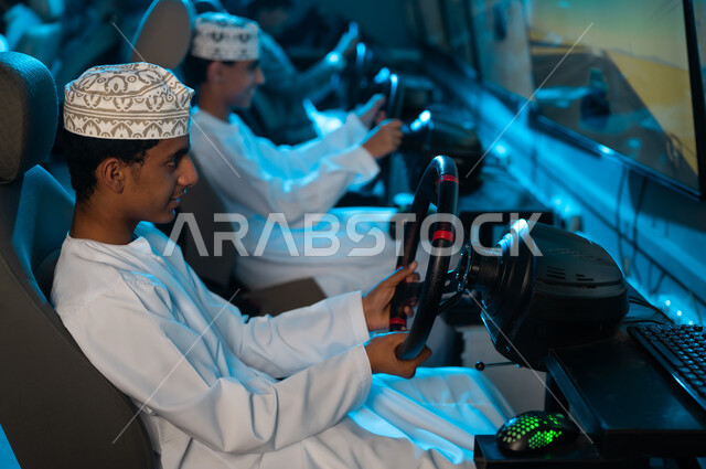شابان عربيان خليجيان عمانيان يرتديان الدشداشة البيضاء و الكمة المطرزة يمسكون عجلة القيادة , لعبة سباق السيارات المثيرة في مركز الالعاب الالكترونية , نشاط ممتع بين الاصدقاء