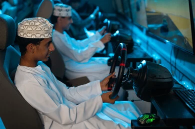 شابان عربيان خليجيان عمانيان يرتديان الدشداشة البيضاء و الكمة المطرزة يمسكون عجلة القيادة , لعبة سباق السيارات المثيرة في مركز الالعاب الالكترونية , نشاط ممتع بين الاصدقاء