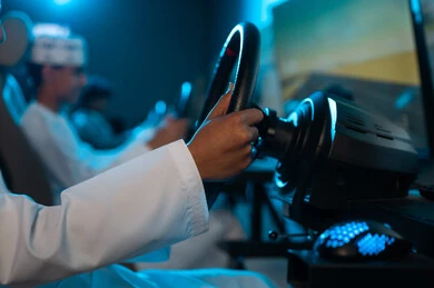 امساك عجلة القيادة بقوة , شاب عربي خليجي عماني يرتدي دشداشة بيضاء يجلس على مقعد السائق , لعبة جماعية تنافسية داخل مركز الالعاب الكترونية , صورة مقربة