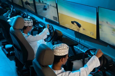 النظرالي الشاشات بتركيز للفوز في سباق السيارات , مجموعة من الشباب العربي الخليجي العماني يرتدي الدشداشة البيضاء و الكمة المطرزة يجلسون على مقعد السائق , العاب ممتعة واجواء شبابية