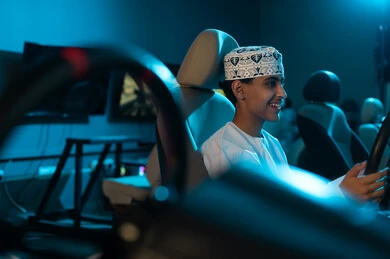 Looking at the screen hanging on the wall while playing, a young Arab Gulf Omani man wearing a white dishdasha and an embroidered kumma sits in the driver's seat, spending special moments, close-up shot.