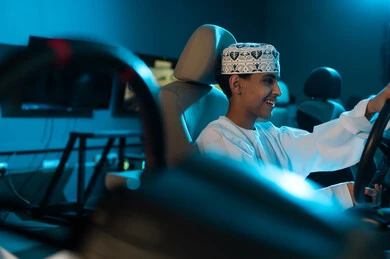 الالعاب المسلية في مراكز الالعاب الالكترونية , شاب عربي خليجي عماني يرتدي الدشداشة البيضاء و الكمة المطرزة , الامساك بعجلة القيادة بثبات , ايماءات السعادة و الابتسامة , صورة مقربة