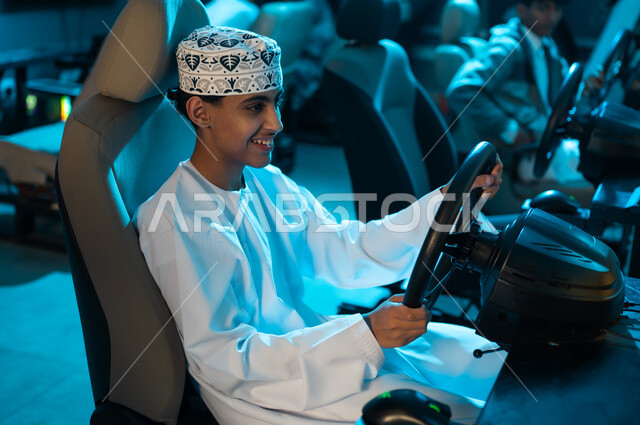 قيادة السياارة بسعادة و فرحة , شاب عربي خليجي عماني صغير يرتدي دشداشة بيضاء و كمة مطرزة يجلس على كرسي لعبة سباق السيارات , انشطة شبابية ممتعة