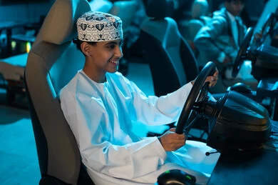 Driving the car with happiness and joy, a young Arab Gulf Omani boy wearing a white dishdasha and an embroidered kumma sits on a racing car game chair, fun youth activities.
