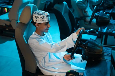لعبة محاكاة حقيقية لسباق السيارات , شاب عربي خليجي عماني يرتدي الدشداشة البيضاء و الكمة المطرزة يجلس على كرسي السائق , اوقات ممتعة في مركز الالعاب الالكترونية , صورة جانبية