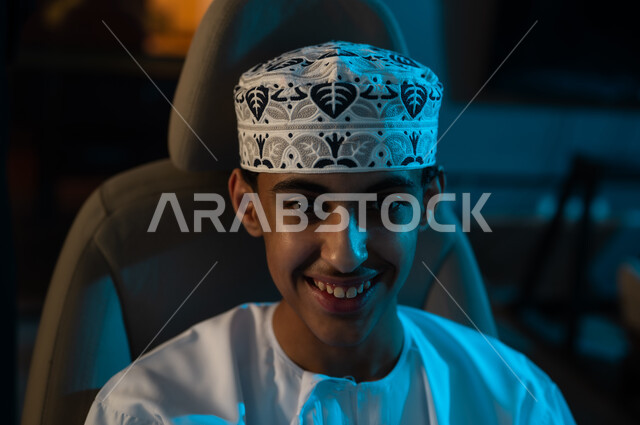 شاب عربي خليجي عماني صغير يرتدي دشداشة بيضاء و كمة مطرزة يجلس على كرسي لعبة سباق السيارات , صورة مقربة , ايماءات السعادة , النظر للكاميرا
