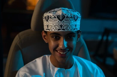 شاب عربي خليجي عماني صغير يرتدي دشداشة بيضاء و كمة مطرزة يجلس على كرسي لعبة سباق السيارات , صورة مقربة , ايماءات السعادة , النظر للكاميرا