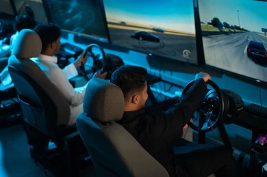 صورة خلفية لرجلان عربيان خليجيان سعوديان يرتديان الثوب الابيض و الاسود يتشاركان في لعبة سباق السيارات , محاكاة للقيادة الحقيقية , الجلوس امام الشاشات المعلقة , ايماءات الحماس و السعادة