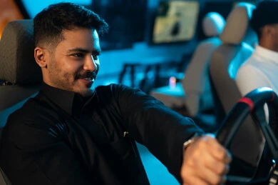 توحية محرك اللعبة يميناً و يساراً باحترافية عالية للفوز في السباق , رجل عربي خليجي سعودي يرتدي الثوب الاسود يجلس خلف مقود لعبة السيارات , لعبة حماسية ممتعة داخل مركز الالعاب الالكترونية