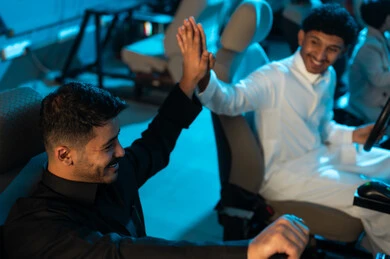Two young Arab Gulf Saudis wearing white and black thobes are sitting behind the steering wheel of a car game, raising their hands in a high five as a sign of joy for winning, looking at the game screen.