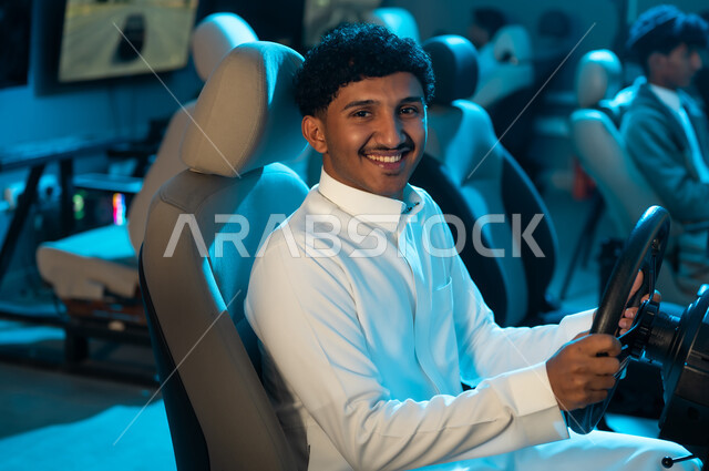 النظر الي الكاميرا بابتسامة , شاب عربي خليجي سعودي يرتدي الثوب الابيض يجلس على كرسي السيارة , لعبة محاكاة سباق السيارات الحقيقية , الاوقات الممتعة داخل مركز الالعاب الالكترونية