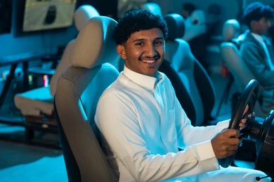 النظر الي الكاميرا بابتسامة , شاب عربي خليجي سعودي يرتدي الثوب الابيض يجلس على كرسي السيارة , لعبة محاكاة سباق السيارات الحقيقية , الاوقات الممتعة داخل مركز الالعاب الالكترونية