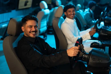 Looking at the camera with a smile, two Arab men from the Gulf, wearing white and black thobes, close-up image, sitting on chairs behind the steering wheel of the car, a simulation game for real driving.