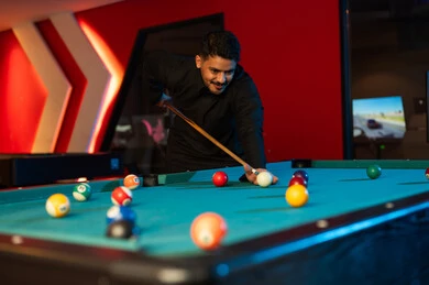 شاب عربي خليجي سعودي يرتدي الثوب الاسود يوجه العصا للكرة البيضاء , قضاء الاوقات المسلية داخل مراكز الالعاب , كرات صغيرة ملونة على طاولة البلياردو