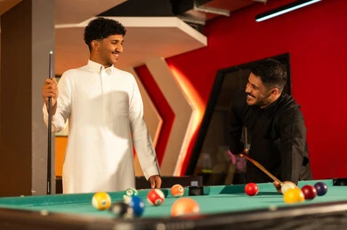 رجلان عربيان خلجيان سعوديان ينظران لبعضهما بسعادة اثناء لعب البلياردو , ايماءات الابتسامة و الفرحة , توجيه العصا الخشبية للكرة البيضاء الصغيرة , الوقوف امام الطاولة الخضراء